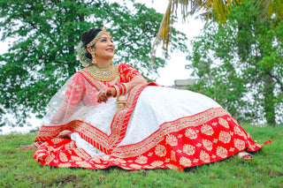 Beautiful Indian bride posing gracefully in a red and white traditional lehenga in a lush outdoor setting.