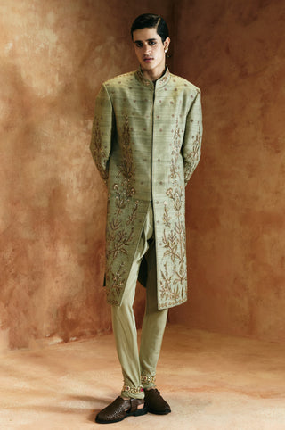 Full-length front view of the green sherwani set with hands behind the back and tonal fitted pants by Aman Takyar at Mrs. G