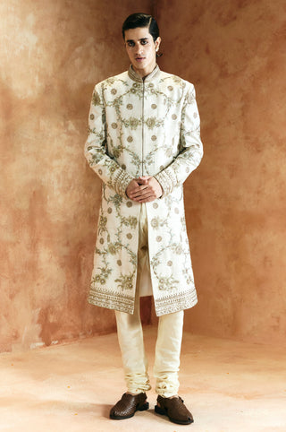 Front view of the ivory sherwani set, hands clasped, with gold and sage vine embroidery by Aman Takyar at Mrs. G