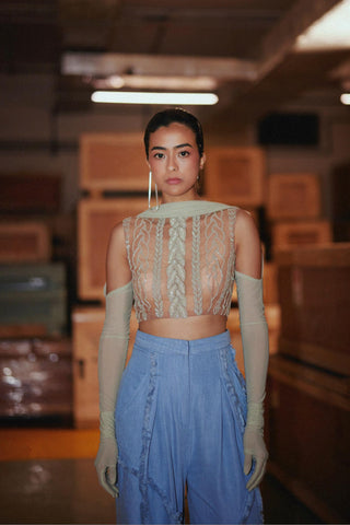A model stands in a dimly lit warehouse or studio space wearing a beige lace and mesh crop top with matching long gloves and light blue high-waisted wide-leg pants.