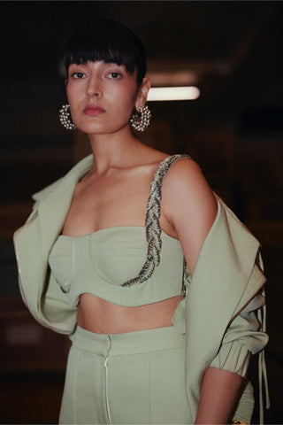 A model wearing a sage green two-piece set consisting of a strapless crop top with a snakeskin-patterned shoulder strap detail and matching high-waisted trousers, styled with a matching draped jacket.