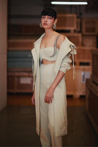 A model stands in a warehouse-like setting wearing a light cream-colored fashion outfit consisting of a long open-front coat, a cropped strapless top with silver chain detailing, and matching trousers.