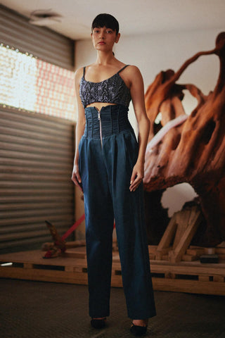 A model stands in a warehouse-like setting wearing a dark blue lace-trimmed crop top and matching high-waisted wide-leg trousers with a front zipper.