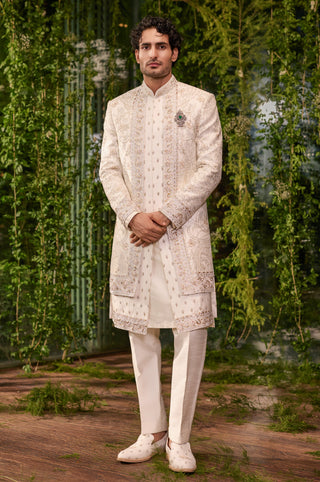 Full-length front view of the ivory bespoke sherwani set with aari embroidery, brooch and ivory trousers by Jigar and Nikita at Mrs. G
