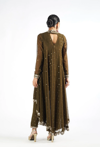 A woman viewed from the back wearing a long brown embroidered dress with sheer sleeves, a high open back, and decorative metallic embellishments, standing against a plain white studio background.