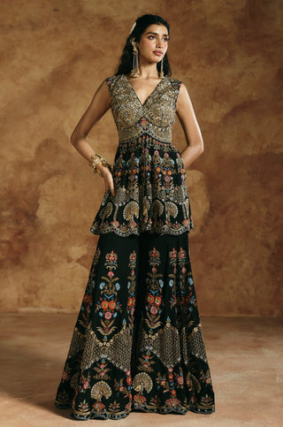 Full-length view with both hands on hips and floral hem embroidery on the black sharara by Aman Takyar at Mrs. G