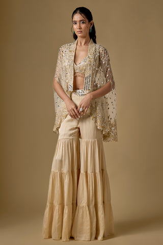 Full-length front view of the beige short mirror cape with a gold hem over a bralette blouse and tiered satin sharara by Asaga at Mrs. G